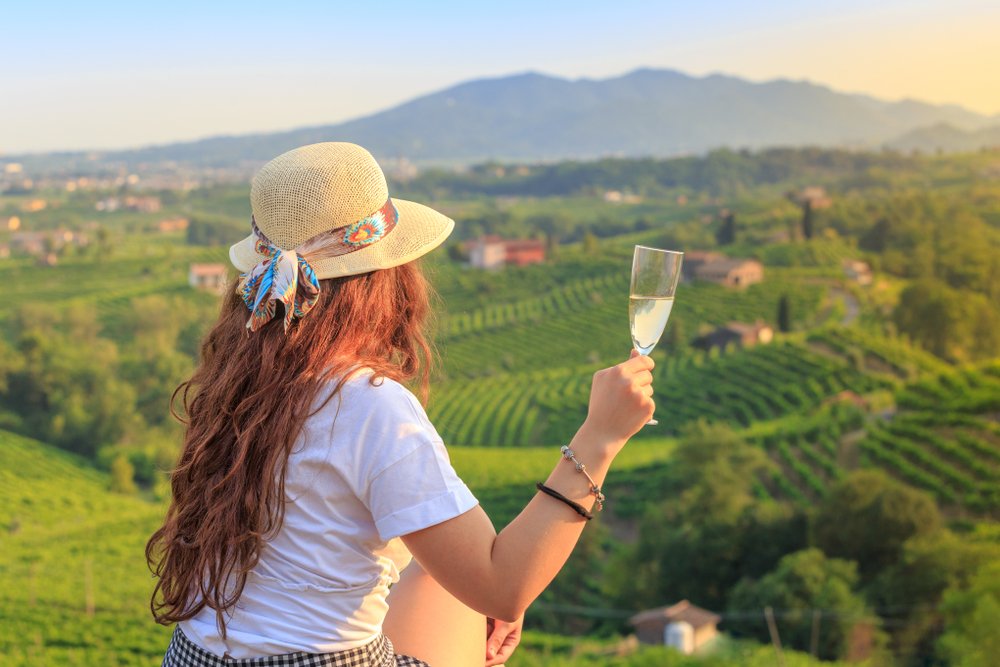 woman in a hat, white shirt and shorts drinking prosecco while looking over vineyards as the sun sets