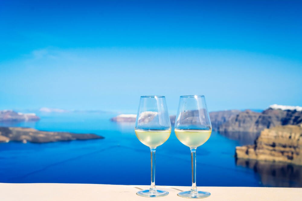 two glasses of white wine against a sunny backdrop of santorini caldera with cliffside views