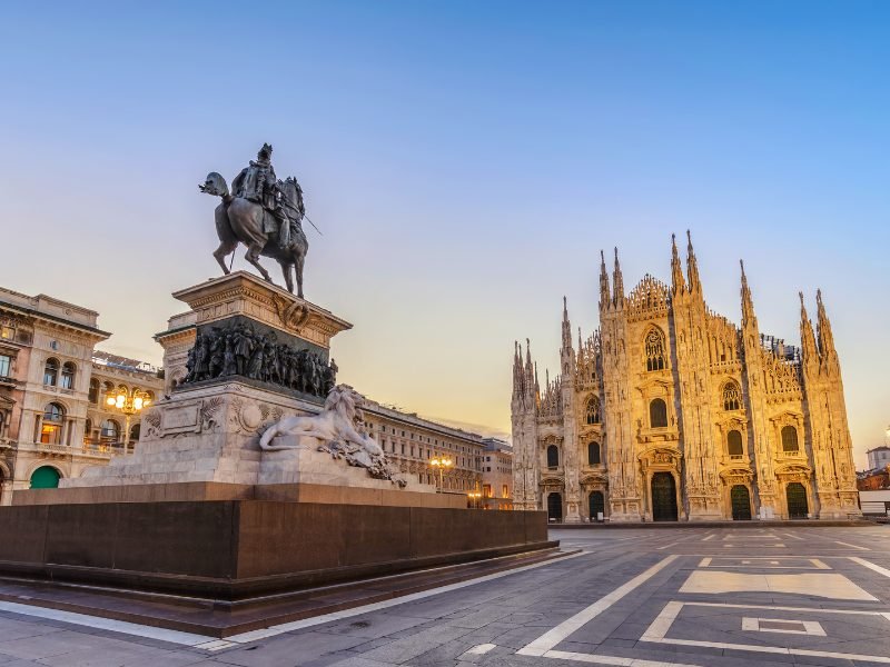 A statue of a man on horseback with a sword, facing the Milan duomo, which is aglow in golden light at either sunrise or sunset
