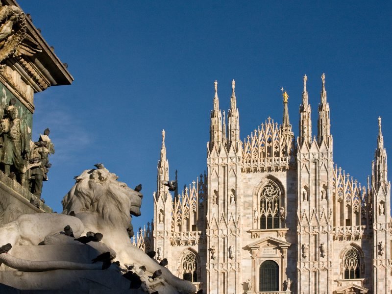 The statue of a lion and the facade of the Milan Duomo church, with the golden madonnina sculpture perched high on one of the spires.