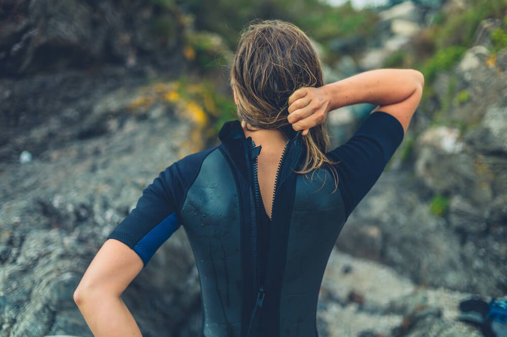 Woman taking off a wetsuit with one hand and holding her hair out the way with another hand. Wearing a shorty wetsuit.