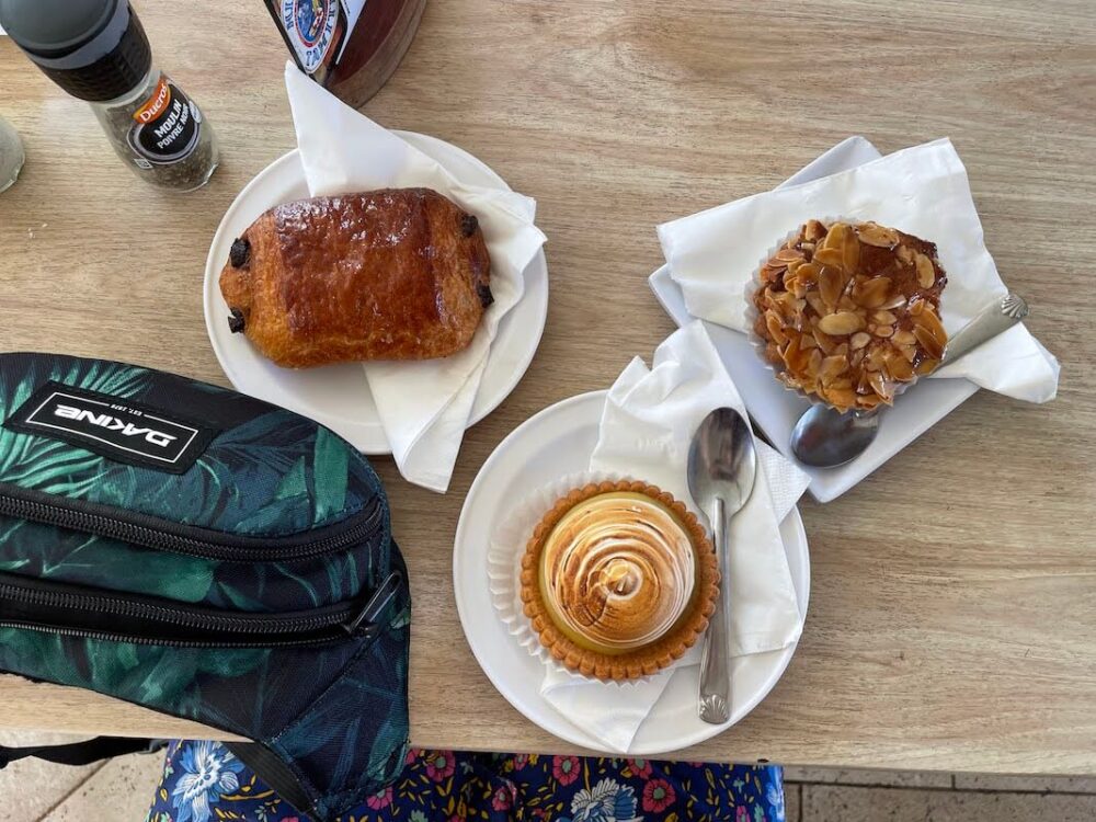 Lemon meringue tart, almond tart an pain au chocolat on a table with a fanny pack on the table, flatlay style