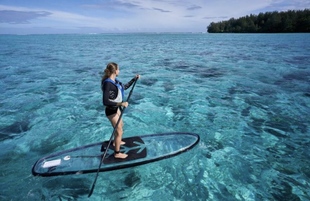 A woman standing on a transparent paddleboard with electric blue clear water beneath her while enjoying a day out