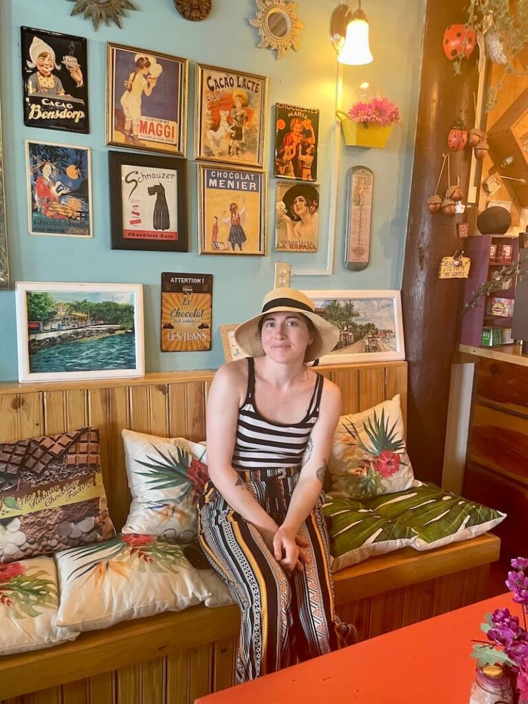 Allison Green wearing a striped tank top, hat, and colorful pants sitting on a colorful bench in the cafe at the roatan chocolate factory