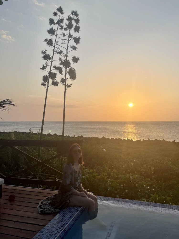 The article author, allison green, wearing a beach cover up with her feet in the pool, smiling as the sun sets behind her in roatan, honduras