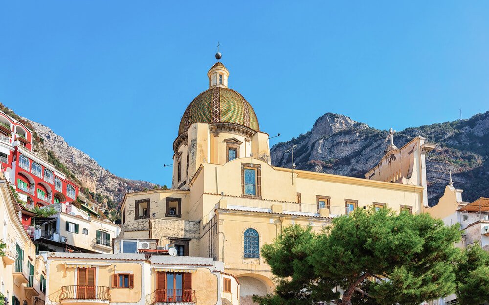 Cityscape and landscape with Santa Maria Assunta Church in Positano town on Amalfi Coast, church has a ceramic tiled dome and sky is blue and cliffs are in the background