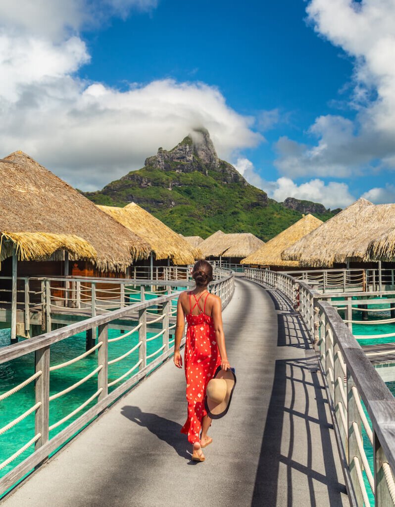 A woman with her hair in a bun, in a red and white dress with a sunhat in hand, walking on the boardwalk of an overwater bungalow hotel in Bora Bora