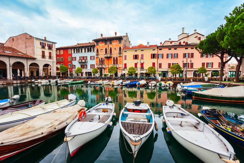 Desenzano del Garda harbor, small boats reflecting in the water, red, blue, orange and yellow buildings on the lakefront harbor area of the town where you start your lake garda itinerary