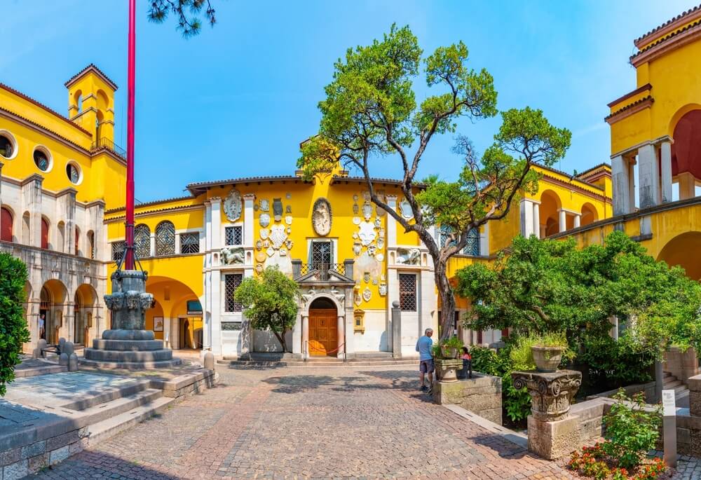 Vittoriale degli italiani palace at Gardone Riviera in Italy, with bright yellow architecture and gardens with a few people in the frame on a sunny day visiting the town of gardone riviera as part of a lake garda itinerary