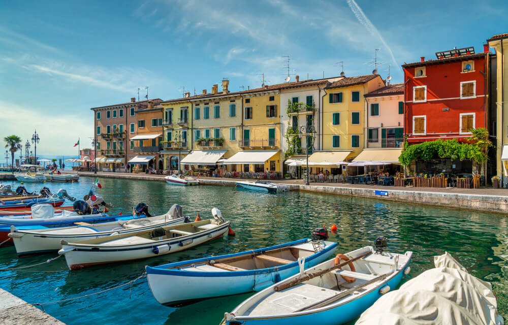 The picturesque town of Lazise on Lake Garda, with red, yellow, lime-green, orange, pink buildings, and small motor-powered boats moored in the town's small marina harbor area