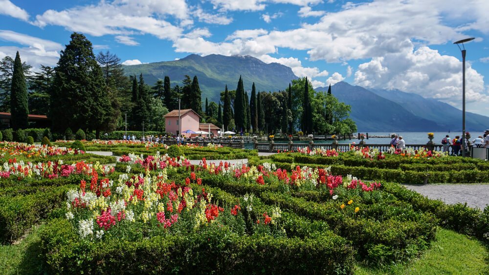 colorful springtime flowerbed in Riva del Garda, on the lake front, with one pink bbuiling visible, people along the walkway
