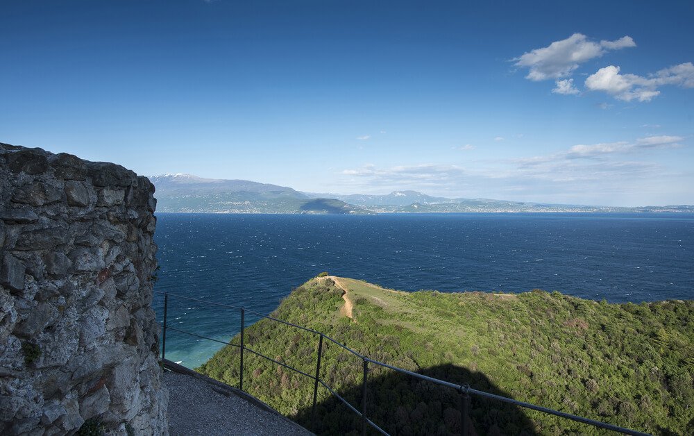 view of Lake Garda from Manerba Rock, with hill and trail with view of lake and towns along the lake around the edge of the lake shore, and some rock formation as part of the park