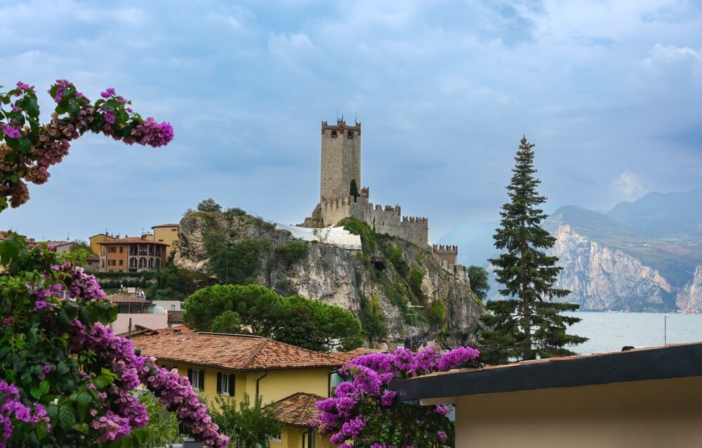 view of the castle in malcesine with purple flowers, yellow and orange buildings, trees, and lake shore on a partly cloudy day