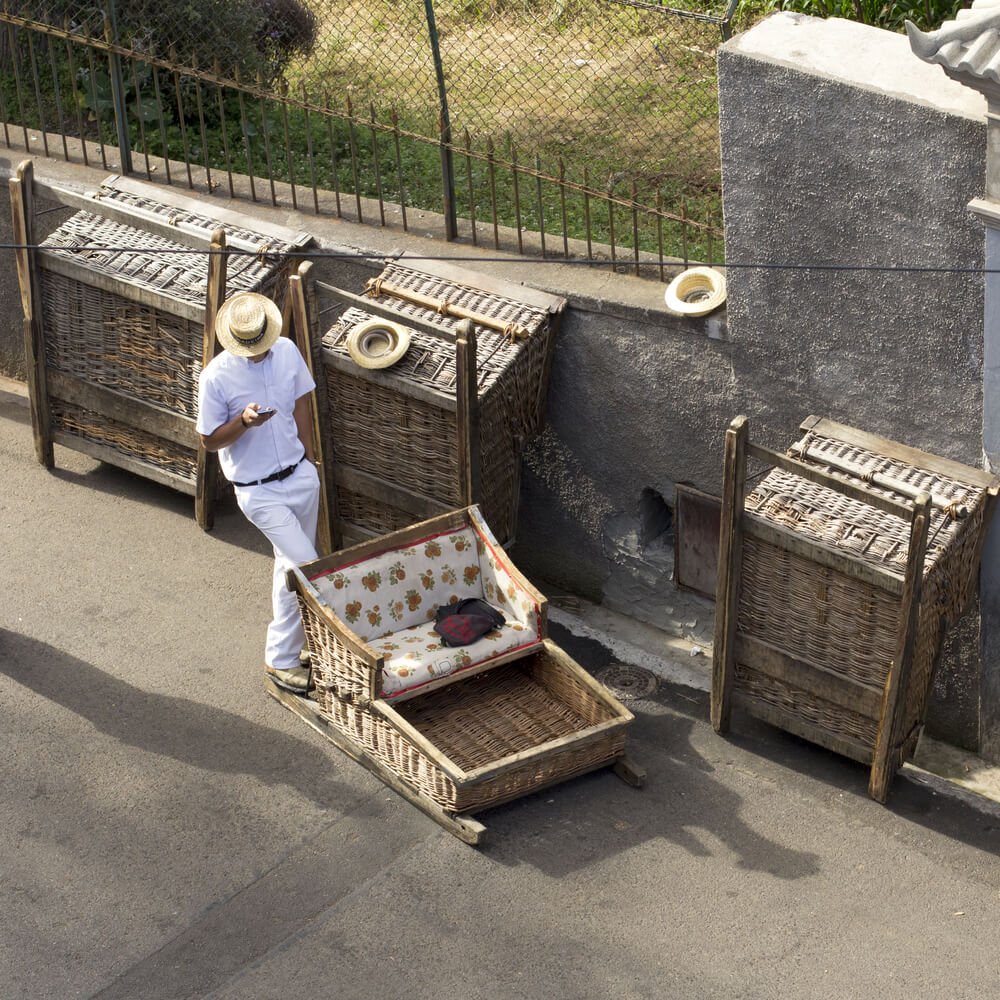 man wearing hat looking at his phone before taking guests on a wicker toboggan ride through madeira