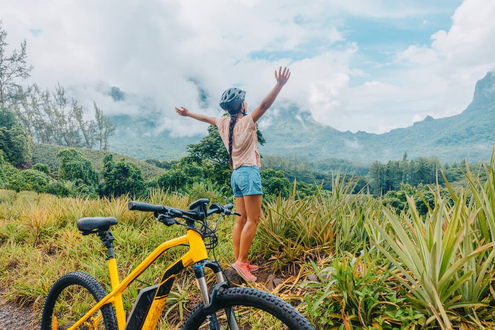 Woman with a helmet on wearing a short sleeve shirt and shorts holding her arms in the arm in a Moorea landscape with a yellow electric bike