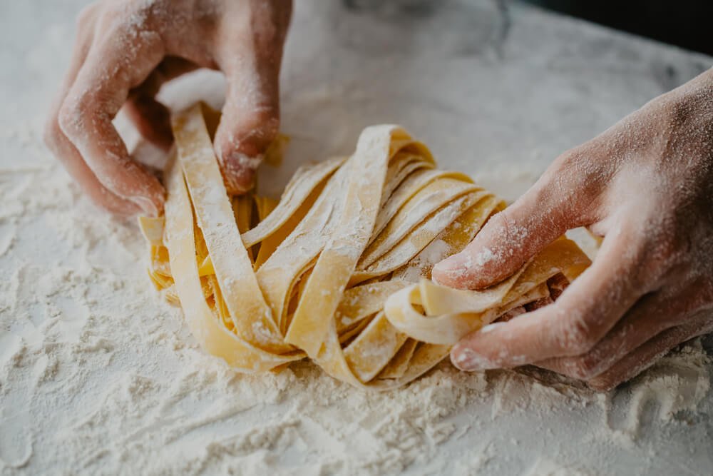 two hands holding fresh made egg pasta that is cut into tagliatelle on a work surface with flour