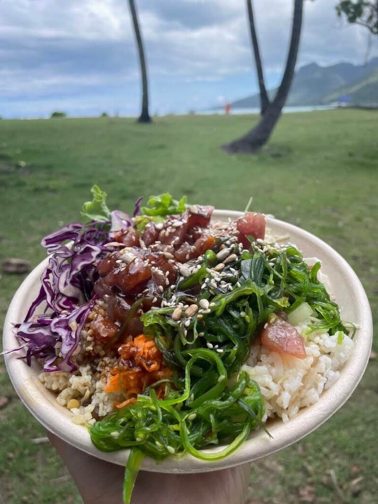 Allison's hand holding a poke bowl in front of the beach in Temae