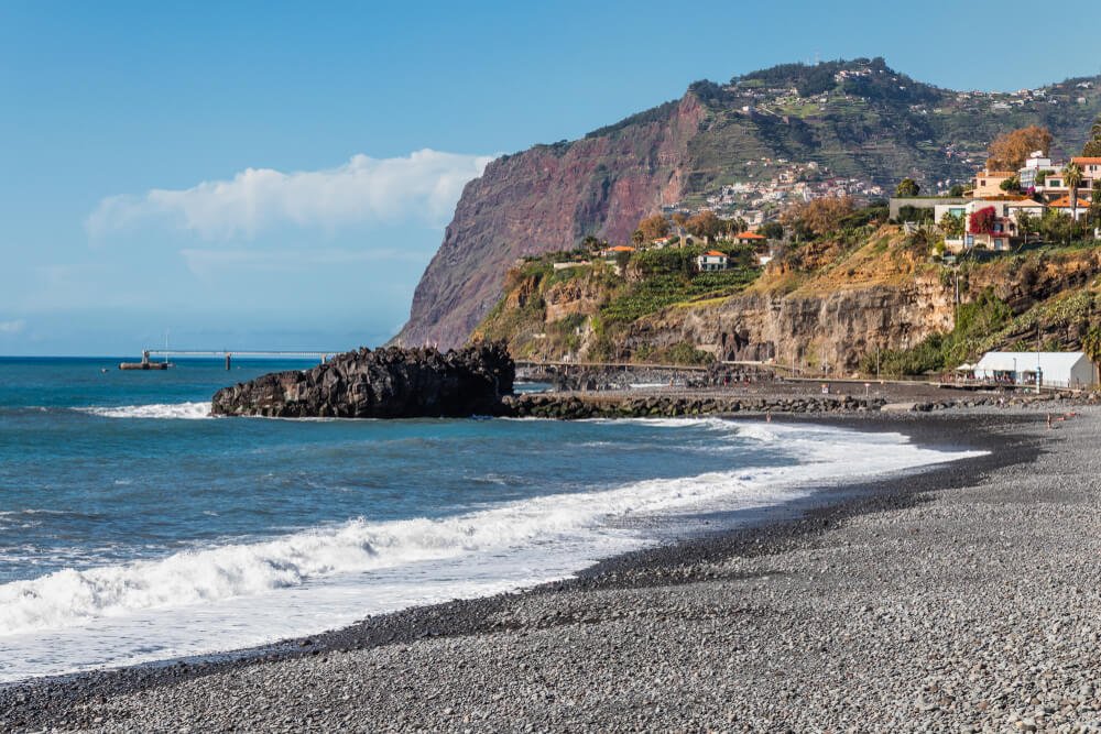 view of the pebble beach of praia formosa in madeira with cliff and hill town behind it