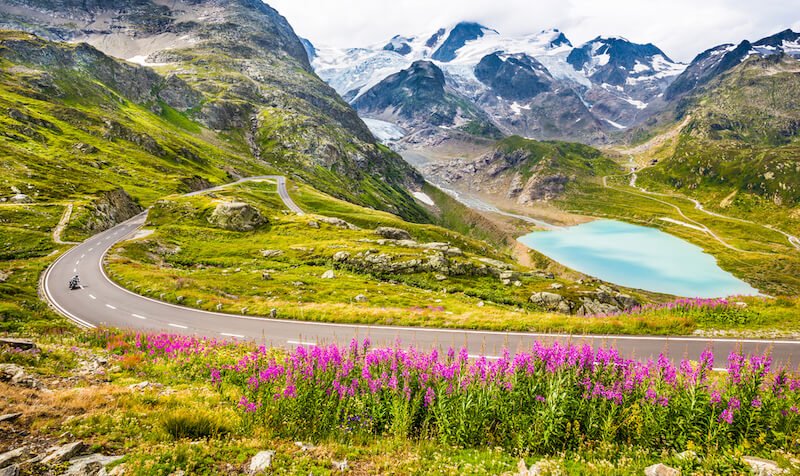 Beautiful view of motorcyclist driving on winding mountain pass road in the Alps through gorgeous scenery with mountain tops, ice glaciers, lakes and green pastures with blooming flowers in summer