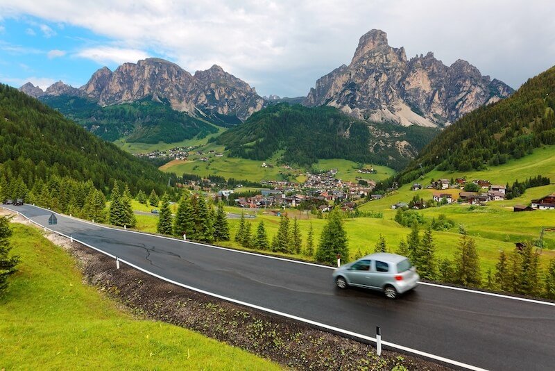 Summer scenery of cars driving on a highway thru a beautiful valley in Dolomiti with villages scattered on the green grassy hillsides of rugged mountains