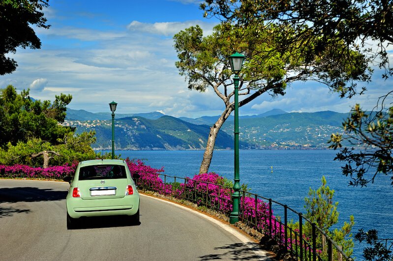 Beautiful road to Portofino from Santa Margherita, pretty pale green car on the road with lots of pink flowers on the side of the road, and lake or sea views.