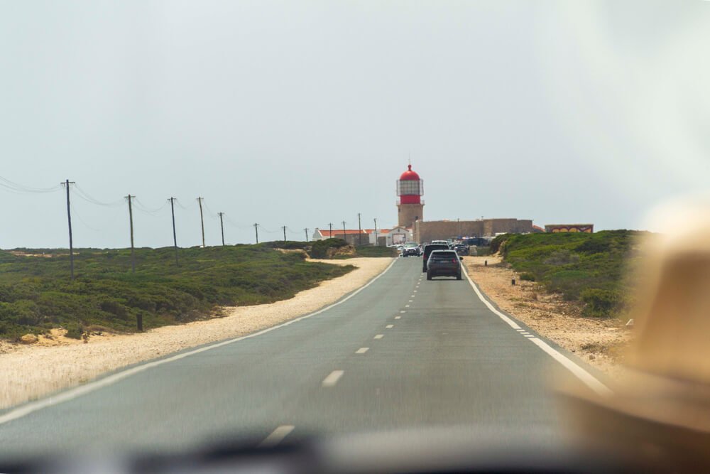 driving in a car in portugal, person with hat blurry passenger, lighthouse visible at the end of road