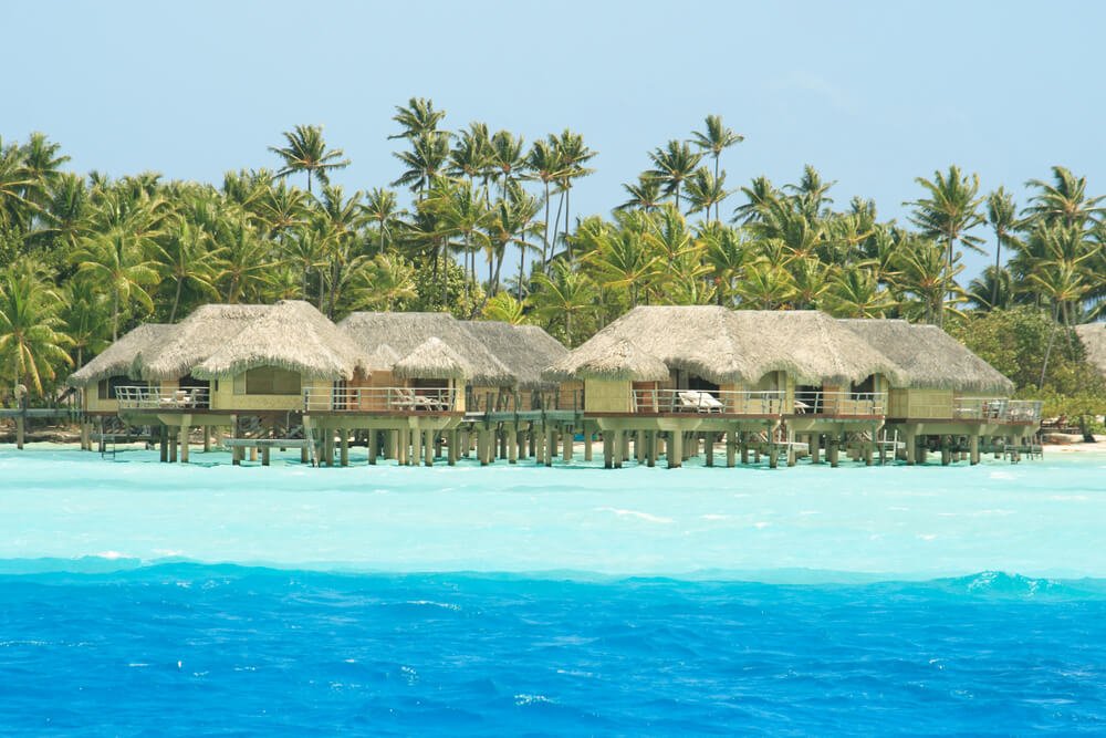 Overwater bungalows of Tahaa with dark blue and light blue water and coconut grove island behind the bungalows