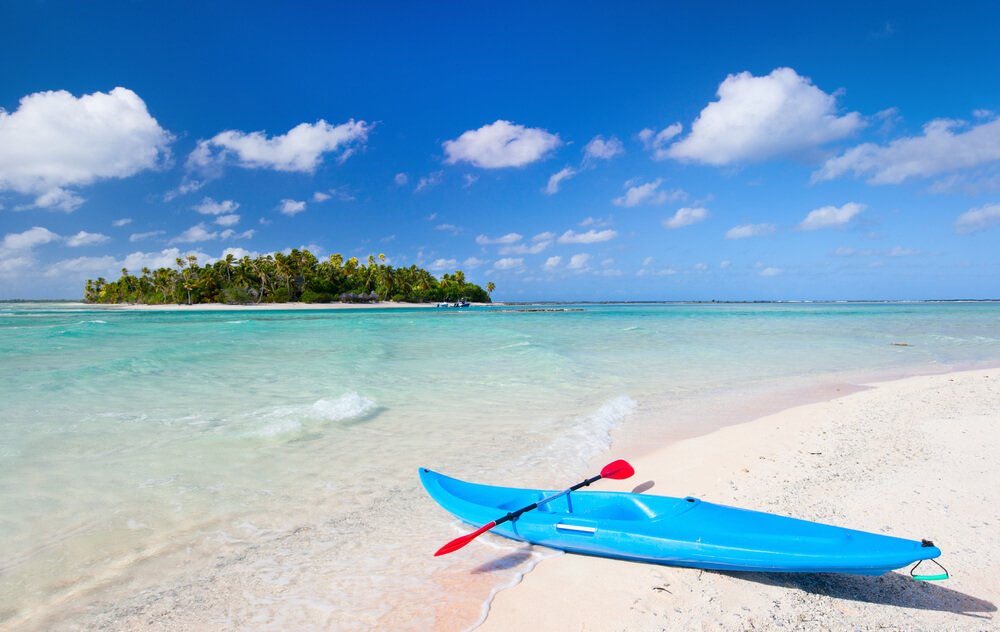 blue kayak with red oar and turquoise waters on a pink sand beach with the palm-covered island out in the water