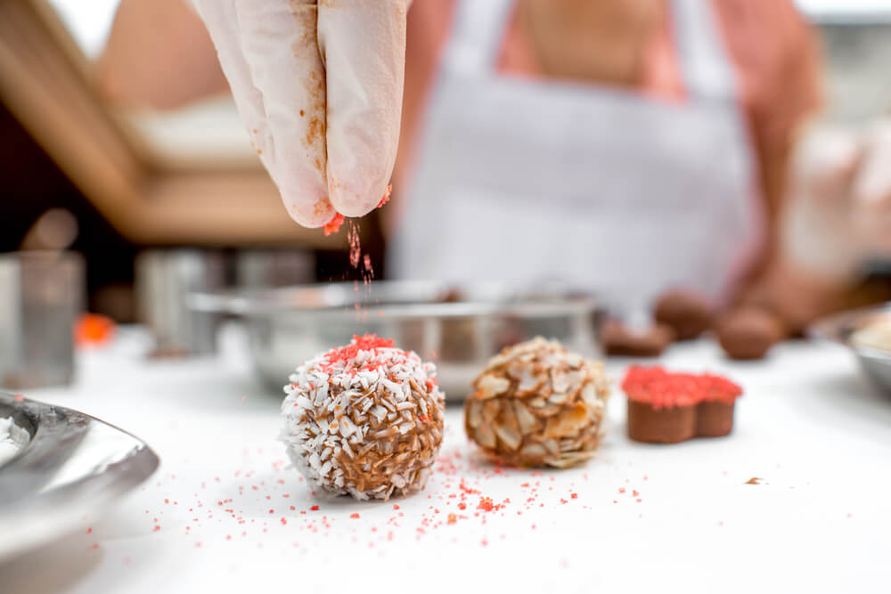 Young woman making and decorating chocolate candy with coconade, red sugar and almonds on the white table
