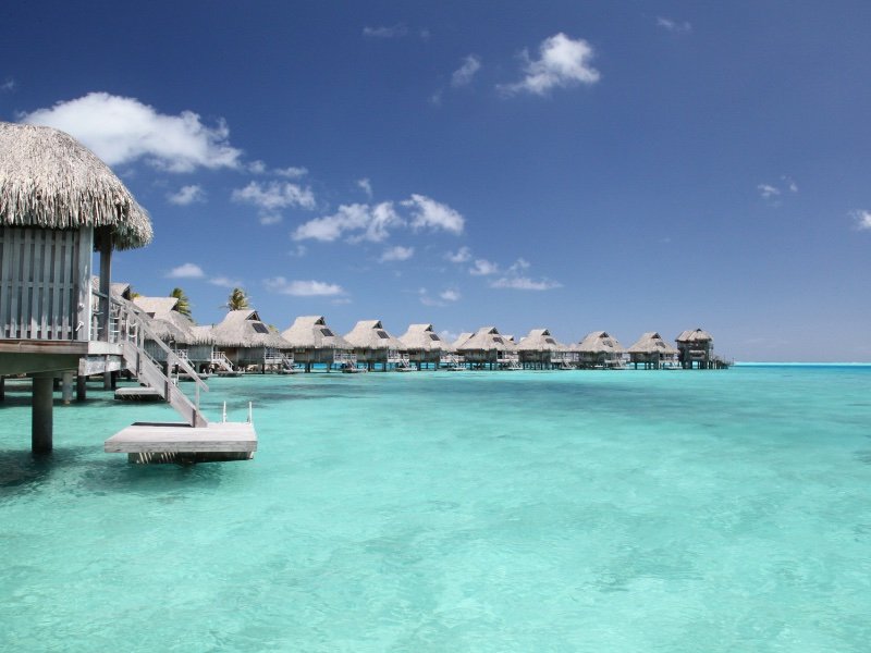 view of the overwater bungalows of bora bora with light blue clear water on a sunny day just a few clouds in the sky