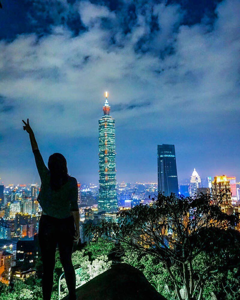 Allison Green standing on a boulder on elephant mountain overlooking the Taipei 101 building at night, her hand up in the air with a silhouette of a peace sign