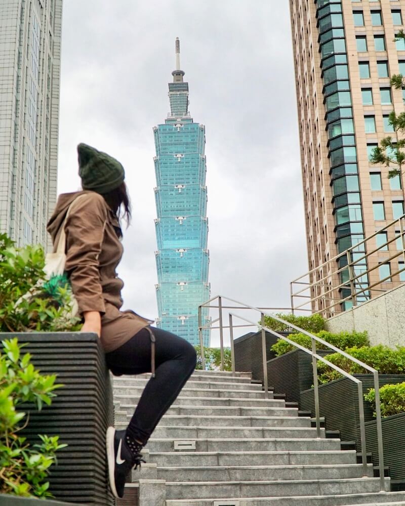Allison Green in a hat, wearing sneakers and a coat, looking at the Taipei 101 in the distance