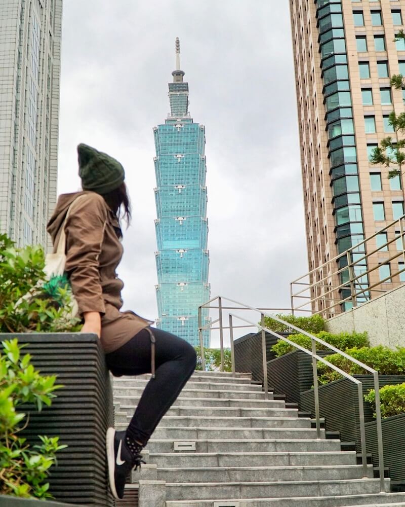 Allison Green wearing an army green jacket and hat and sneakers,  looking at the architecture of the taipei 101 from afar