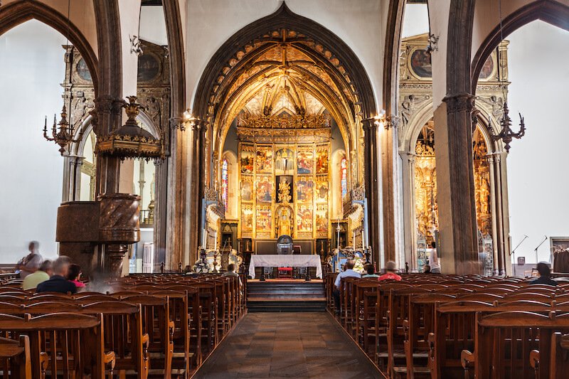 the ornate gilded interior of the funchal catehdral, with lots of altarpieces in the nave, wooden chairs that serve as pews, and otherwise white walls with wooden detail in the parts that are not decorative