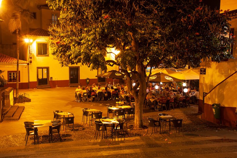 Alley in the old town with restaurants, by night, orange lamplight casting a color over the scene of people eating dinner late at night, Funchal, Madeira