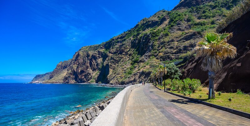 Jardim do Mar village with Promenade at beautiful coast of Madeira island, brilliant blue sea and paved pathway with palm tree and rocky terrain at the end of the path