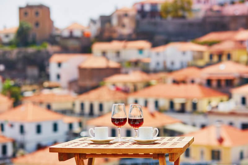 view of two glasses of madeira wine next to cups of coffee, on a table on a terrace, with a view of funchal behind it