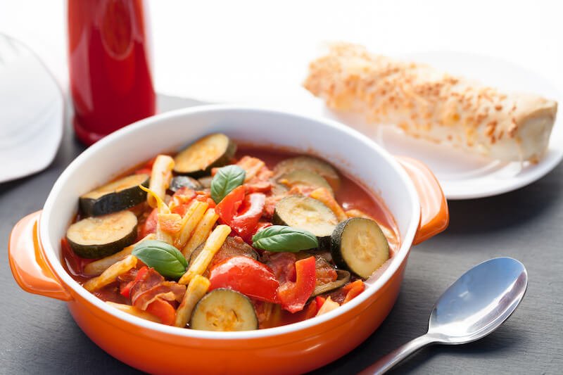 ratatouille with red pepper, tomatoes and green beans, served in an orange cocotte ceramic bowl, with fork, spoon and bread