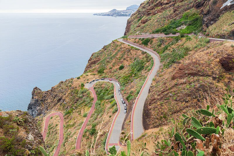 a winding, serpentine road in madeira from the mountains going down to the beachy coastline, with some cars parked along the narrow road