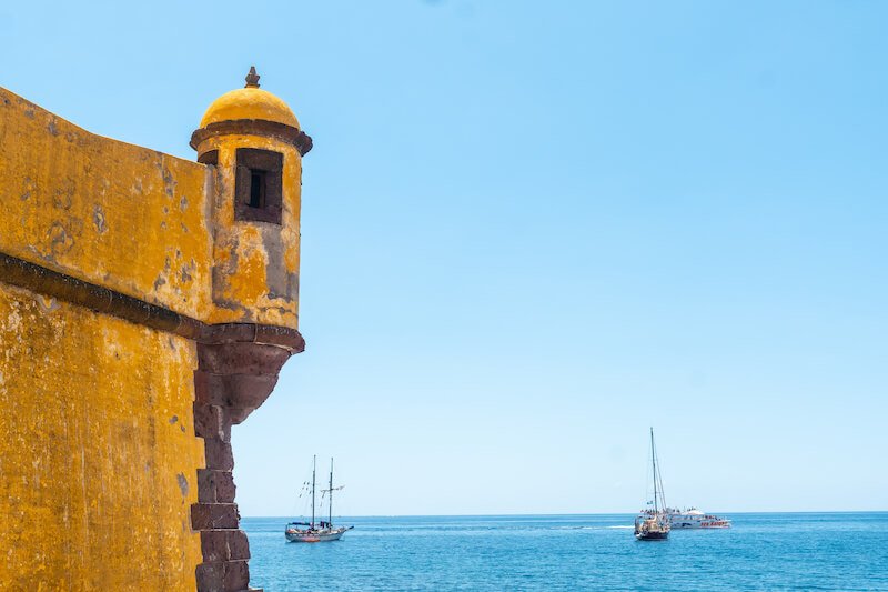 the mustard yellow fort de sao tiago, against the backdrop of azure blue ocean and three sailboats in the distance