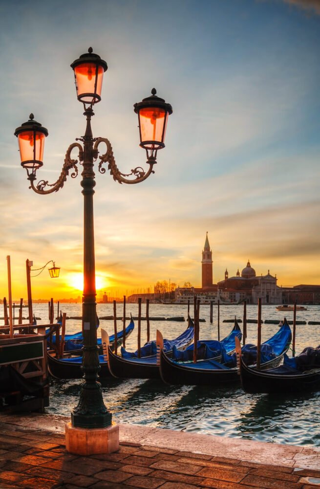 view of gondolas at night with the background of venice skyline in the back and a street lamp with three lanterns