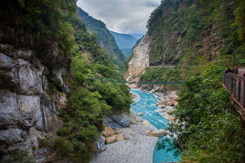 brilliant blue river going down the middle of a gorge with hillsides covered in trees with a suspension bridge