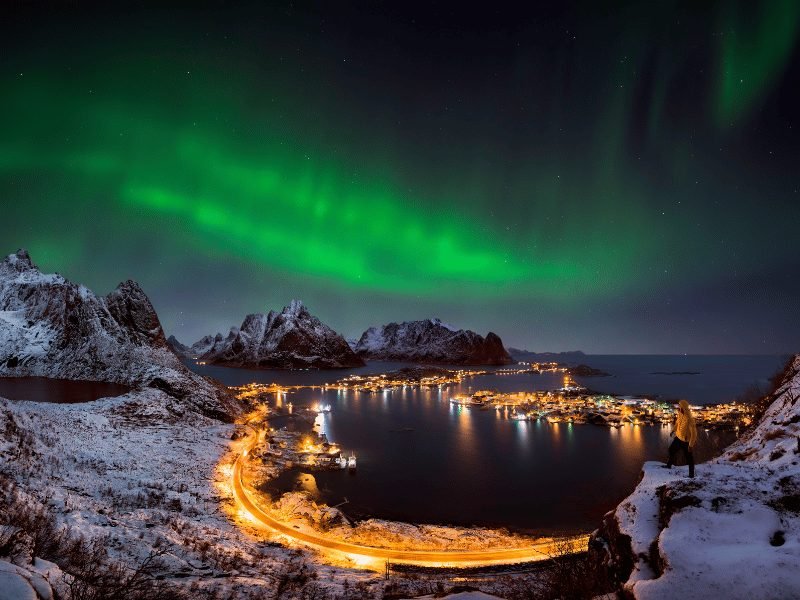 northern lights over lofoten islands in norway with a man in a yellow jacket overlooking all the scenery