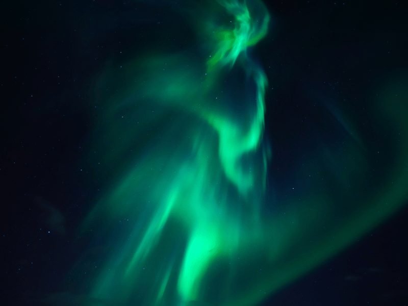 The northern lights in iceland making a spiral up to the sky in bright green and pale blue colors against a midnight-black sky with some stars