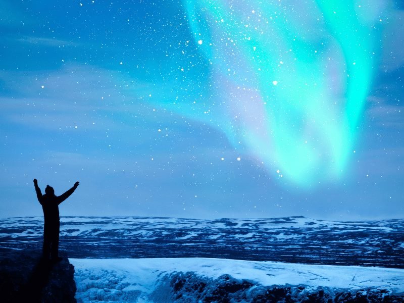 person wearing a hat and jacket and putting their arms up in the sky as the aurora dances overhead