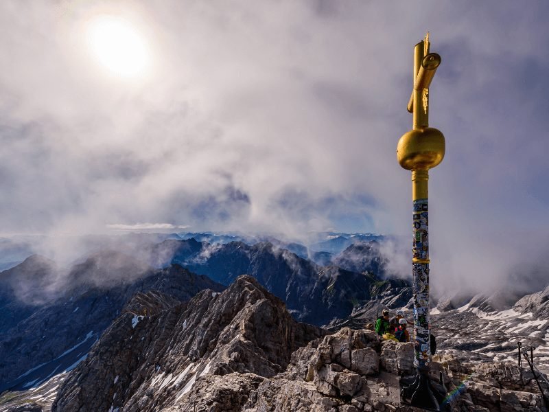 Zugspitze mountain peak with its gold cross that marks the summit and a small group of hikers approaching, the sky is misty and cloudy but you can still see mountains all around