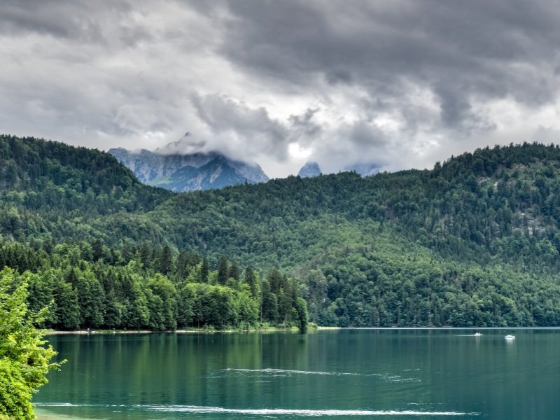 the clear lake waters of alpsee with turquoise water and a few boats and overcast sky with mountain background