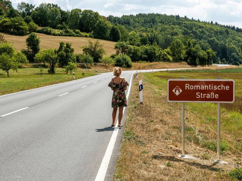 woman in a dress standing by a road sign that says romantische strasse (romantic road) in germany