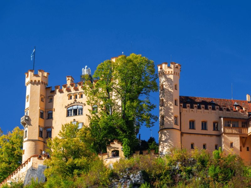 view of Hohenschwangau castle, a pale peach csatle, with summer trees and blue sky