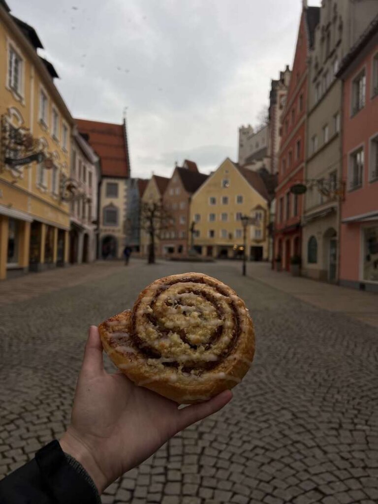 Nicole Westcott's hand holding a Nussschnucken pastry (like a cinnamon roll) with the backdrop of Fussen behind her